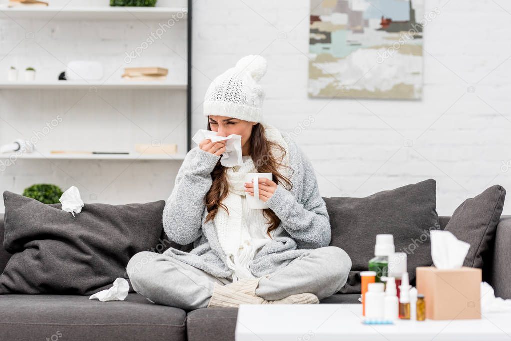 sick young woman in warm clothes sneezing while sitting on couch at home
