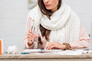 cropped shot of sick young woman in scarf holding glass of water at workplace clipart