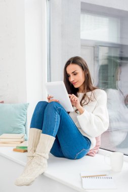 güzel genç kadın evde yastıkları ile pencere üzerinde tablet kullanma
