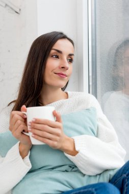 beautiful young woman with cup of coffee looking through window at home clipart
