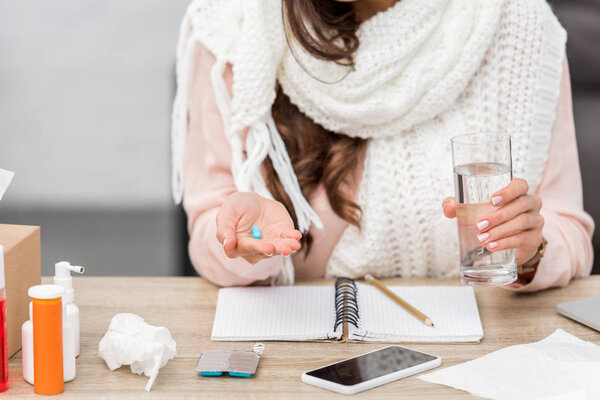 cropped shot of sick young woman in scarf holding glass of water and pill at workplace