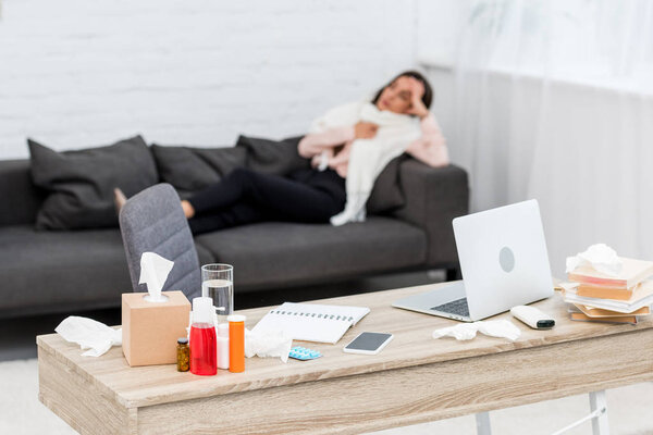 sick young woman lying on couch with work desk full of medicines on foreground 