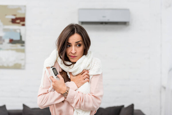 freezed young woman in scarf holding remote control with air conditioner on background