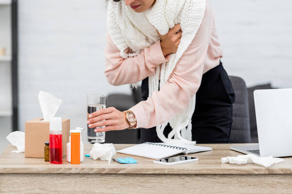 cropped shot of suffering sick young businesswoman taking glass of water from desk with meds