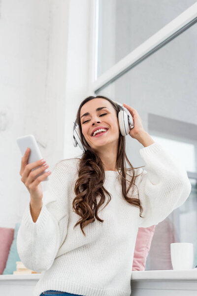 вид снизу на улыбающуюся молодую женщину, слушающую музыку со смартфона и беспроводных наушников дома
