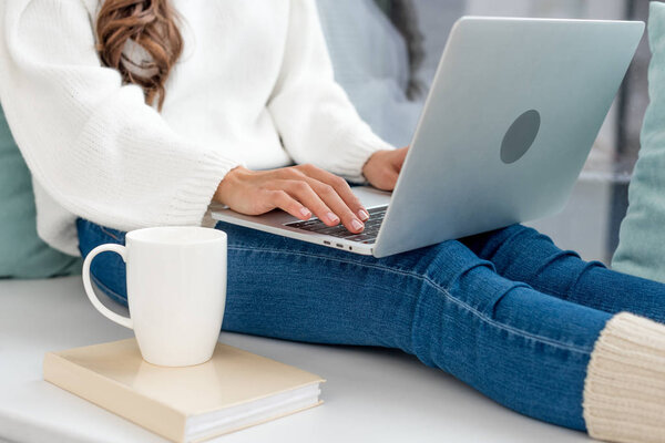 cropped shot of freelancer with cup of coffee working with laptop on windowsill at home