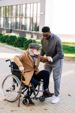 genç Afro-Amerikan hemşire yardımcı kıdemli adam sokakta tekerlekli sandalye almak için devre dışı.