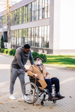 senior disabled man in wheelchair with walking stick and african american cuidador talking on street clipart