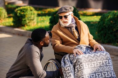 senior disabled man in wheelchair and african american nurse chatting on street clipart