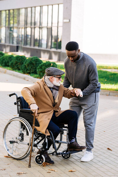 young african american nurse helping senior disabled man to get up from wheelchair on street