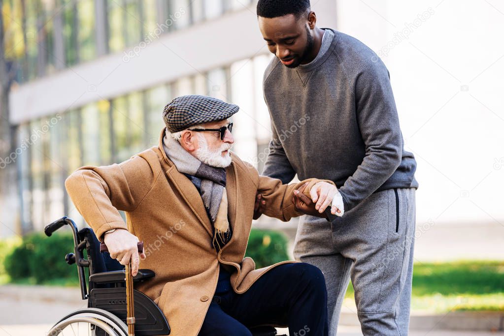 young african american man helping senior disabled man to get up from wheelchair on street