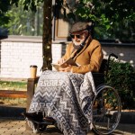 Idoso deficiente homem em cadeira de rodas com xícara de papel de livro de leitura de café na rua