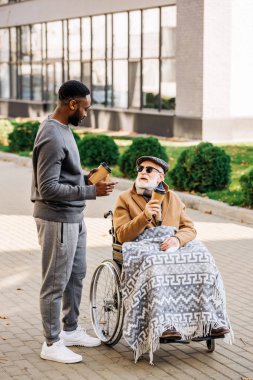 Kıdemli adam ekose ile tekerlekli sandalyede ve Afro-Amerikan erkek tome sokak kağıt bardak kahve ile birlikte harcama devre dışı
