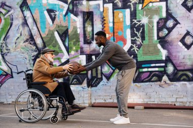 side view of senior disabled man in wheelchair and african american man playing basketball together on street clipart