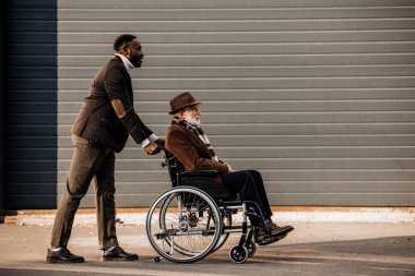 side view of senior disabled man in wheelchair and african american man riding by street clipart