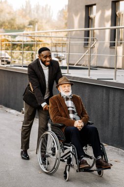 happy senior disabled man in wheelchair and african american man riding by street clipart