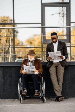 adam tekerlekli sandalyede ve Afro-Amerikan yakışıklı Gazeteler birlikte sokakta okuma kıdemli devre dışı