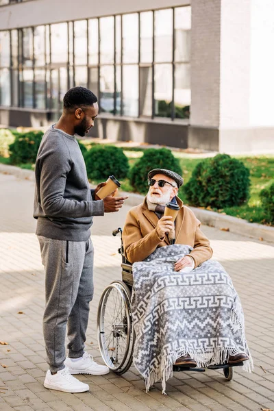 senior disabled man in wheelchair with plaid and african american man spending tome together on street with paper cups of coffee