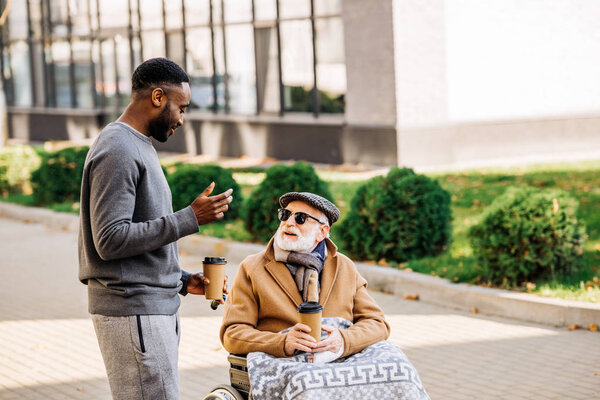 smiling senior disabled man in wheelchair with plaid and african american man spending tome together on street with paper cups of coffee