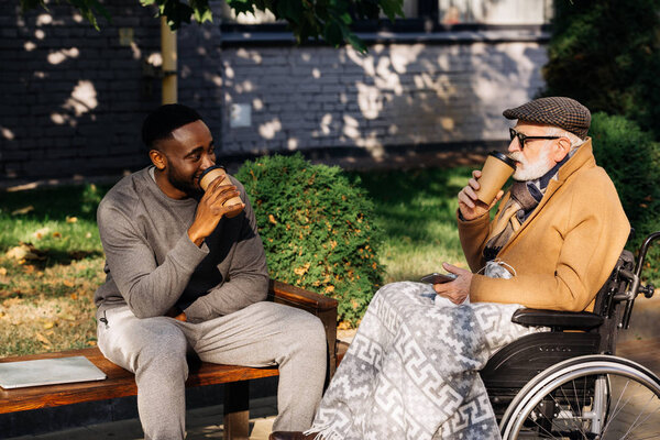 senior disabled man in wheelchair and african american man drinking coffee from paper cups together on street