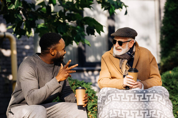 senior disabled man in wheelchair and african american man drinking coffee from paper cups and chatting on street
