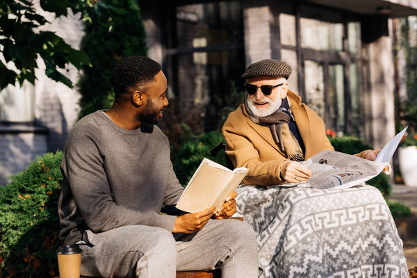 senior disabled man in wheelchair and african american man reading book and newspaper together on street