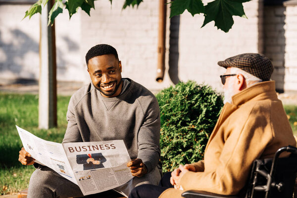 happy senior disabled man in wheelchair and african american man spending time together and reading newspaper on street