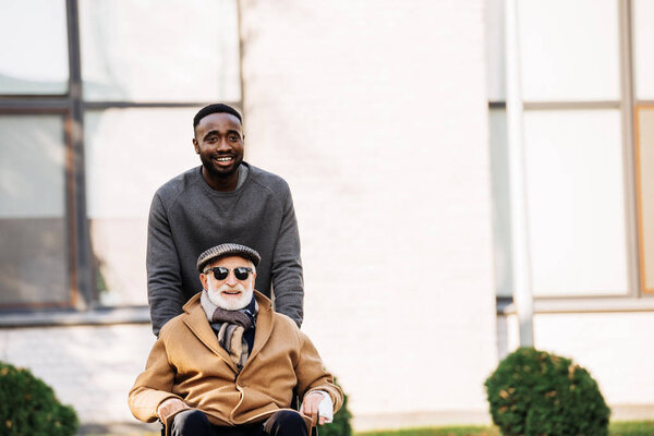 smiling senior disabled man in wheelchair and african american man riding by street together