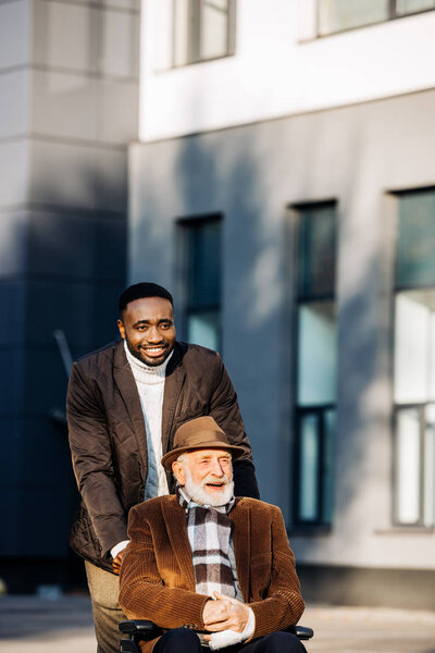 happy senior disabled man in wheelchair and african american cuidador riding by street