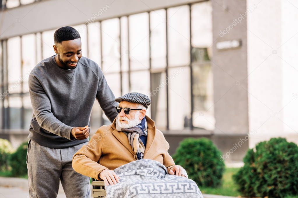 happy african american man giving joint to senior disabled man in wheelchair wjhile they spending time together on street