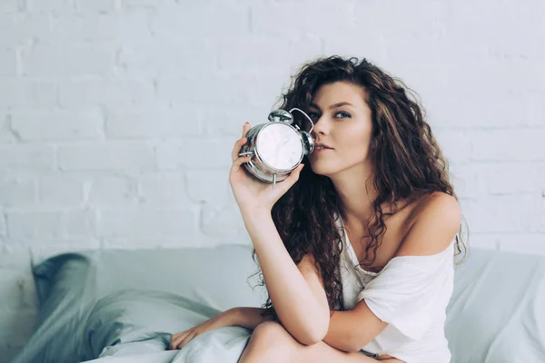 自宅の目覚し時計とベッドの上に座って陽気な巻き毛の若い女性 — ストック写真
