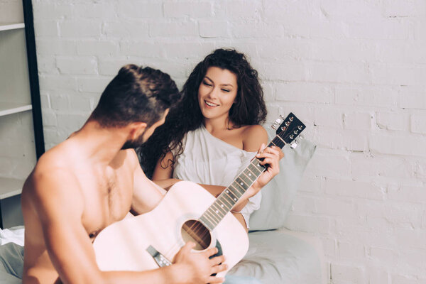 man playing on acoustic guitar while his happy girlfriend sitting near in bedroom at home