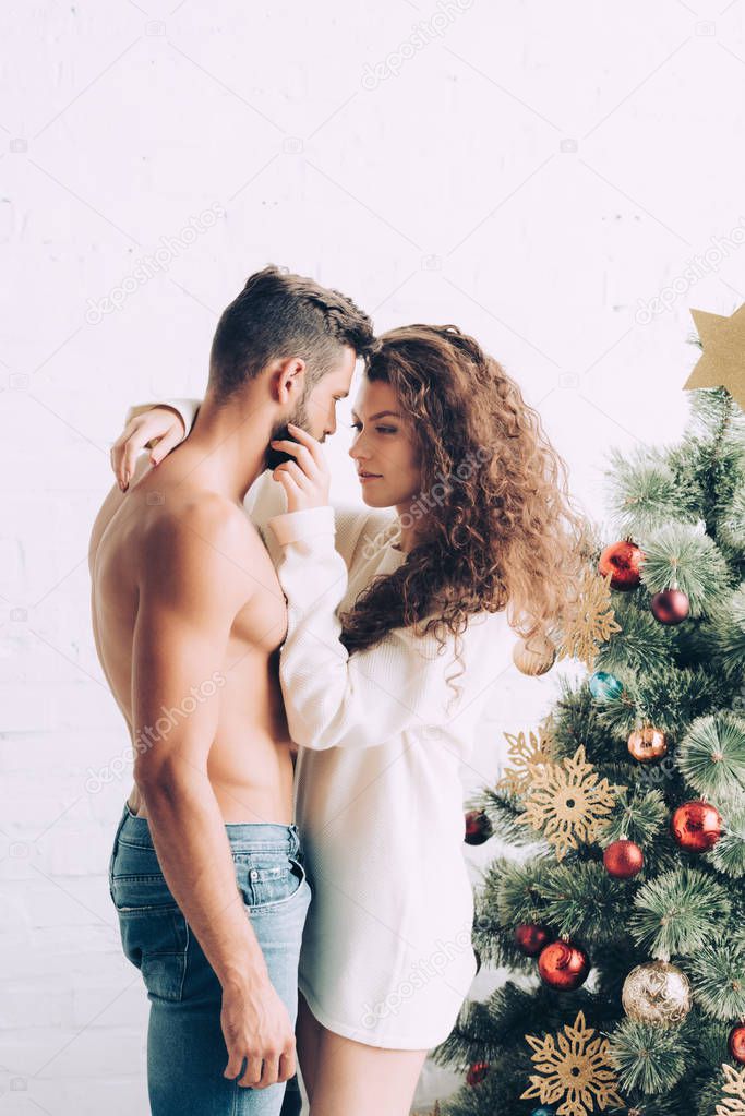 cheerful young woman embracing shirtless boyfriend near christmas tree at home