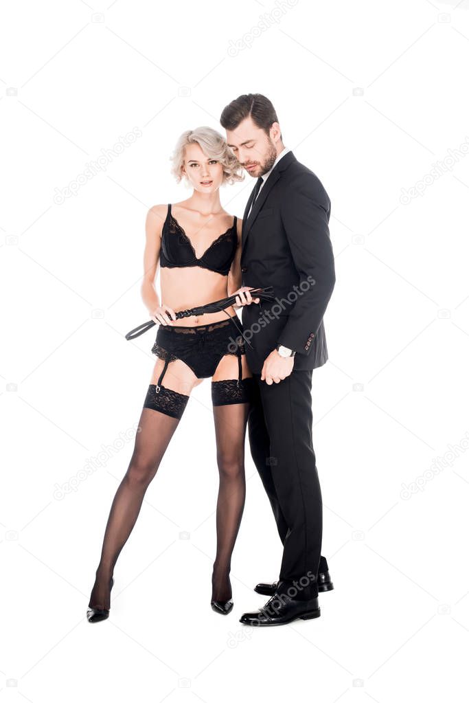 Woman in lingerie holding whip and standing near handsome man in black costume isolated on white
