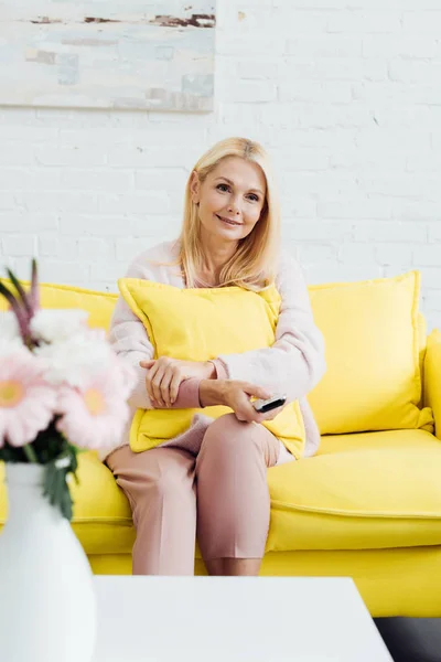 美丽的微笑成熟的女人坐在明亮的黄色沙发上与电视遥控器 — 免费的图库照片