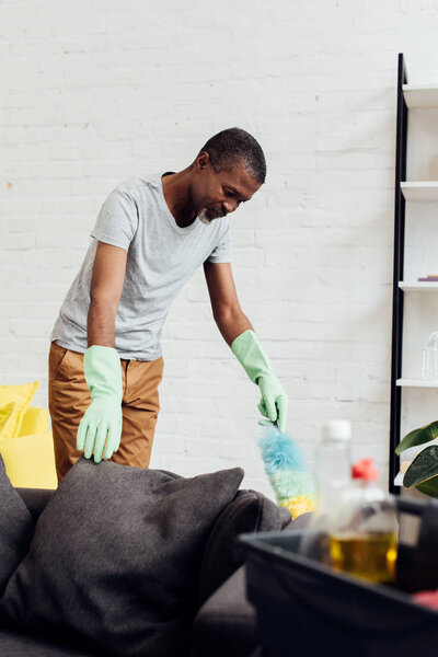 handsome african american man in rubber gloves doing housecleaning with duster