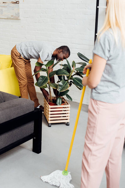 blonde woman holding mop while african american man putting plant in wooden pot