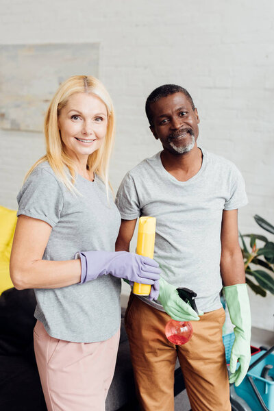 smiling blonde woman and african american man in rubber gloves holding housecleaning equipment 