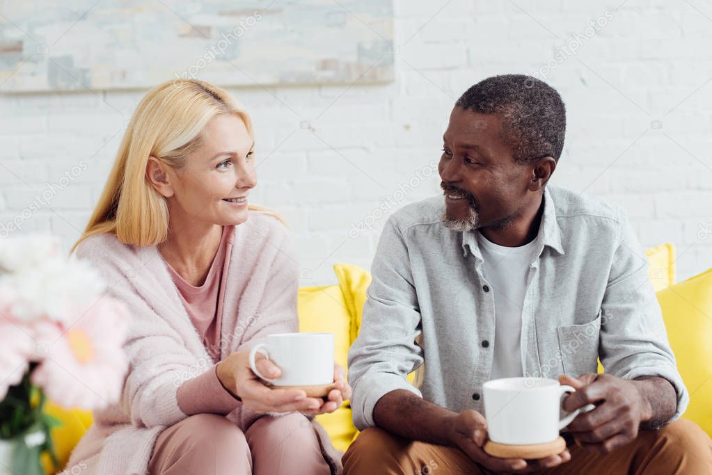 smiling mature woman sitting on sofa with african american man and drinking coffee together