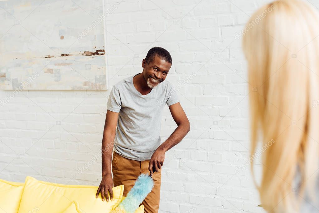 smiling african american man cleaning sofa with duster