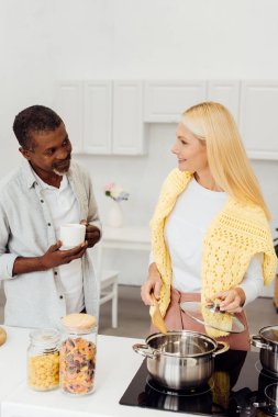 mature blonde woman cooking dinner while african american man drinking coffee clipart