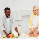 微笑的妇女切西红柿在切菜板上, 而非洲美国男子拿着盘子与蔬菜