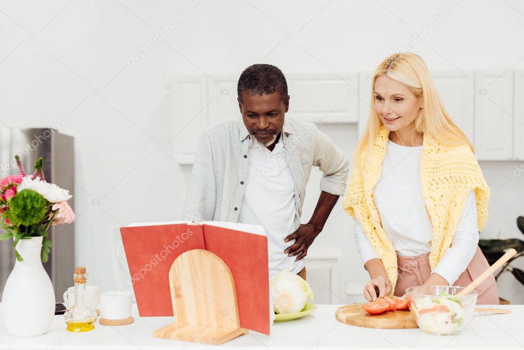 happy interracial couple cooking dinner together and reading recipe in cookbook at kitchen