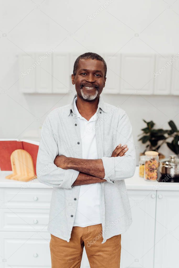 happy african american mature man with arms crossed standing at kitchen