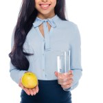 Cropped shot of smiling african american woman holding glass of water and apple isolated on white