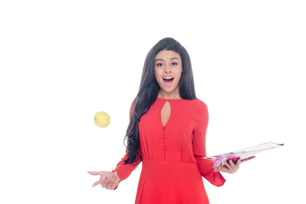 шокированная африканская американка в красном платье держит журнал и бросает яблоко изолированы на белом
