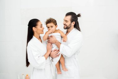 Mother and father looking at toddler son in white towel and smiling  clipart