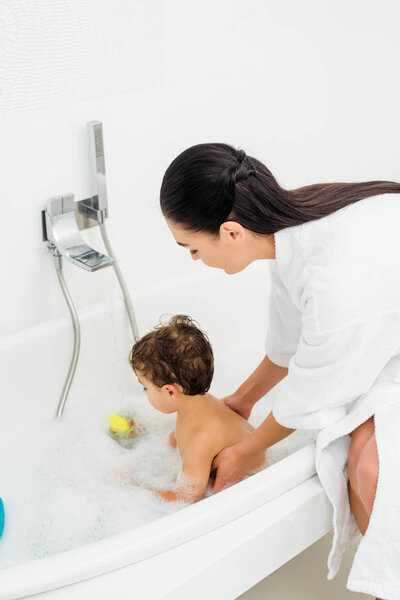 Mom in bathrobe washing son in white bathroom