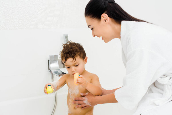 Smiling mother washing son in foam in white bathroom