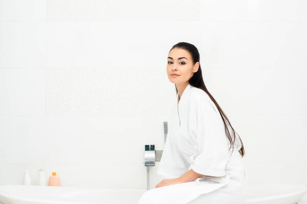 Attractive woman in bathrobe sitting in white bathroom 